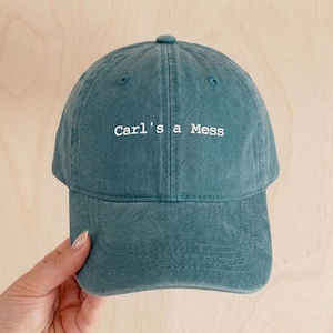 May include: Teal baseball cap with the embroidered text "Carl's a Mess" in white. The cap has a curved brim and a top button. The hat is made of a soft, textured fabric.