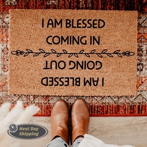 May include: A brown coir doormat with the text "I am blessed coming in I am blessed going out" in black lettering. The text is framed by a decorative line of leaves.