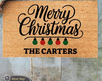 May include: A brown coir doormat with the text "Merry Christmas" in black cursive font. A string of red and green Christmas lights hangs below the text. The text "THE CARTERS" is printed in black below the lights.