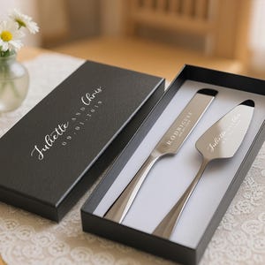May include: A wedding cake knife and server set in a black box. The box is open, revealing the silver utensils with engraved names and a date. The box lid has the names "Juliette and Chris" and the date "09-01-2019".