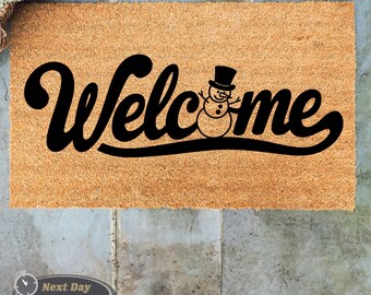 May include: A brown coir doormat with the word "Welcome" in black cursive lettering. A snowman with a black top hat is positioned within the word "Welcome".