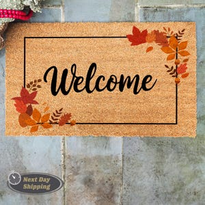 May include: A brown coir doormat with a black frame and the word "Welcome" in black script. The frame is decorated with a cluster of orange, red, and yellow fall leaves in the top right corner.