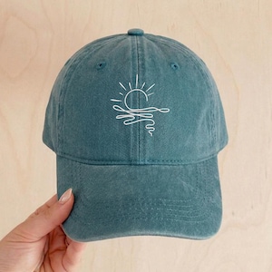 May include: Teal baseball cap with a white embroidered design of a sun over water. The cap has a curved peak and an adjustable strap at the back. The hat is made of a soft, washed cotton material.