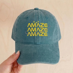 May include: Teal baseball cap with the word "AMAZE" embroidered in yellow. The word is stacked three times with a curved line around the bottom two words. The top of the cap has a small button.
