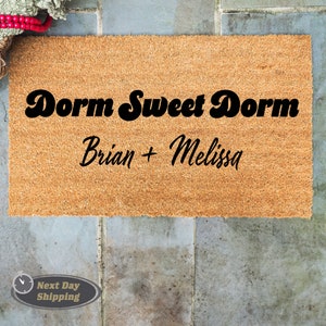 May include: A brown doormat with the text "Dorm Sweet Dorm" in a retro font and the names "Brian + Melissa" written below in a cursive font.