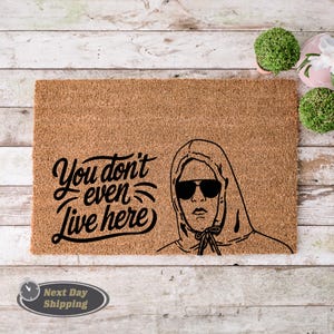 Pode incluir: Um tapete de coco castanho com o texto "You don't even live here" em fonte cursiva preta. Uma ilustra&ccedil;&atilde;o em preto e branco de uma pessoa usando um moletom com capuz e &oacute;culos de sol est&aacute; no lado direito do tapete.