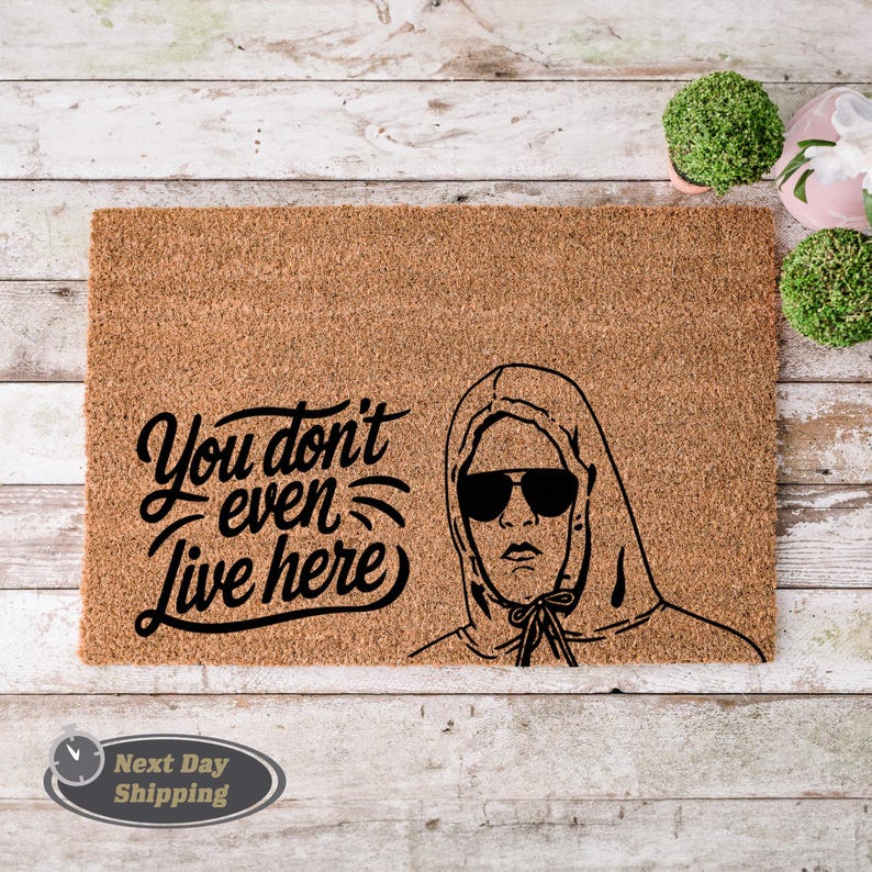 Pode incluir: Um tapete de coco castanho com o texto "You don't even live here" em fonte cursiva preta. Uma ilustra&ccedil;&atilde;o em preto e branco de uma pessoa usando um moletom com capuz e &oacute;culos de sol est&aacute; no lado direito do tapete.