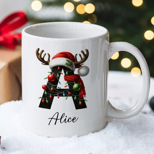 May include: White ceramic mug with a black letter A decorated with Christmas lights, ornaments, and a reindeer wearing a Santa hat. The name "Alice" is written below the letter.