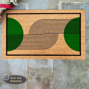 May include: A rectangular doormat with a natural coir fiber background. The design features two green semi-circles and black curved lines. A black border frames the design. The mat is placed on a gray tiled surface.