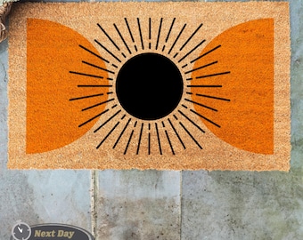 May include: A brown coir doormat with a black sun design and two orange half circles. The sun has rays extending from it.