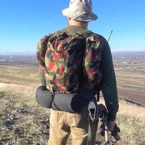 May include: A beige bucket hat, an olive green long-sleeved shirt, tan trousers, and a camouflage backpack with a red, black, and green pattern. A dark gray rolled blanket is attached to the bottom of the pack, and a rifle is held in one hand.