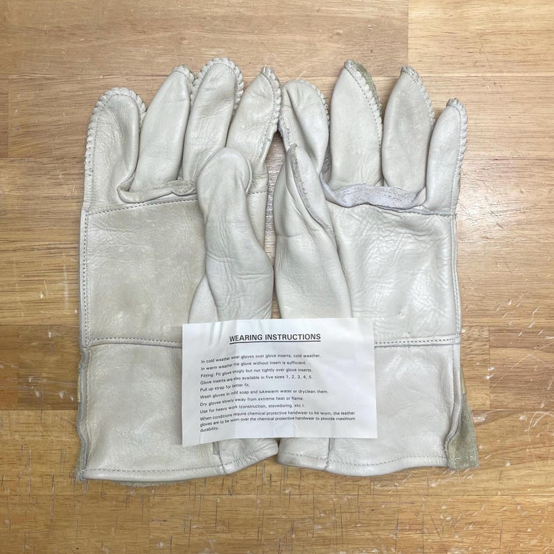 May include: A pair of off-white leather work gloves with reinforced stitching. The gloves have a textured appearance and are laid out on a wooden surface. A paper with wearing instructions is placed between the gloves.