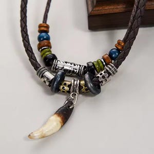 Può includere: Collana in pelle intrecciata marrone con perline decorative blu, verdi e marroni. La collana presenta dettagli in metallo argentato e un ciondolo a forma di dente con una sfumatura dal marrone al crema.