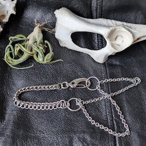 Puede incluir: Una cadena de plata con un cierre de langosta y un anillo. La cadena está sobre una superficie de cuero negro.