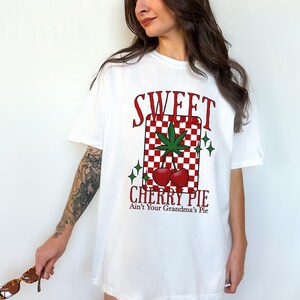May include: White t-shirt with a graphic design featuring the words "SWEET CHERRY PIE" in red, along with a cannabis leaf and cherries. The design is set against a red and white checkered background.