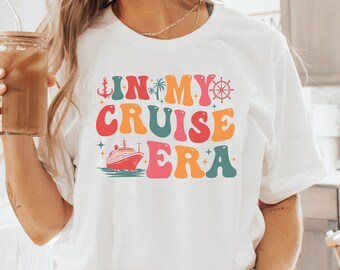 Puede incluir: Una camiseta blanca con el texto "In My Cruise Era" en una fuente retro colorida. El texto incluye un barco de crucero, un ancla, una palmera y un volante.