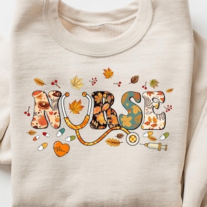 Pode incluir: Uma camisa de moletom bege com a palavra "NURSE" impressa em letras coloridas com um design de outono. As letras s&atilde;o decoradas com folhas, flores e s&iacute;mbolos m&eacute;dicos como um estetosc&oacute;pio e uma seringa.