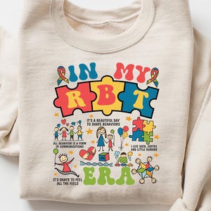 Puede incluir: Una sudadera beige con un diseño gráfico colorido que dice "In My RBT Era". El gráfico presenta piezas de rompecabezas, un arcoíris y figuras de palo.