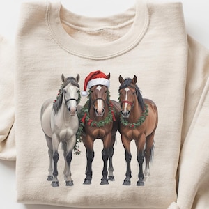 May include: A beige sweatshirt with a Christmas-themed design featuring three horses wearing holly wreaths and a Santa hat. The horses are brown, white, and brown.