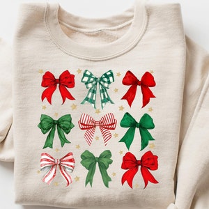 May include: A beige sweatshirt featuring a festive design of red and green bows with gold stars. The bows are arranged in a grid pattern.