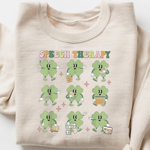 Op de afbeelding: Een beige sweatshirt met de tekst "SPEECH THERAPY" in regenboogkleuren. Het ontwerp bevat negen groene vierbladige klavers met verschillende gezichtsuitdrukkingen en die verschillende voorwerpen vasthouden die te maken hebben met logopedie.