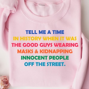 May include: A light pink sweatshirt with the text "TELL ME A TIME IN HISTORY WHEN IT WAS THE GOOD GUYS WEARING MASKS & KIDNAPPING INNOCENT PEOPLE OFF THE STREET." in rainbow colors. The sweatshirt is folded on a white surface with pink flowers.