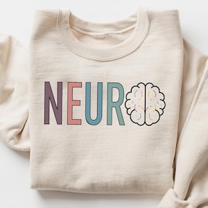 Peut inclure: Un sweat-shirt beige avec le mot "NEURO" en lettres colorées et un dessin de cerveau. Les lettres sont violettes, roses et turquoise. Le dessin du cerveau est souligné en noir avec des couleurs pastel à l'intérieur. Les manches sont retroussées.