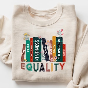 May include: Beige sweatshirt with a colorful graphic of books. Each book spine displays a word like "Love," "Kindness," and "Equality." Floral accents are placed throughout the design. The sweatshirt is folded, with the sleeves casually draped.