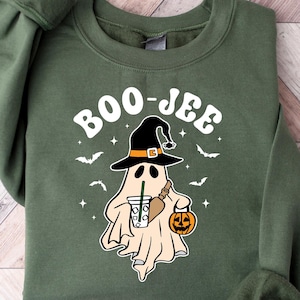 Może przedstawiać: Zielona bluza z nadrukiem białego ducha w czarodziejskim kapeluszu, trzymającego dynię i kubek kawy. Na bluzie widnieje napis "BOO-JEE".