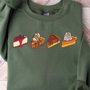 May include: A green sweatshirt with a line of five different pie slices printed on the front. The slices include cheesecake, apple pie, pecan pie, pumpkin pie, and a pie with whipped cream.