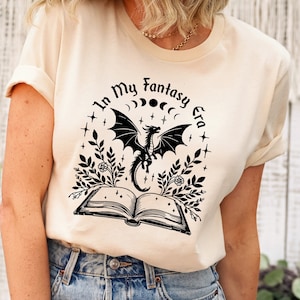以下が含まれることがあります： クリーム色のTシャツ。黒いグラフィックデザインで、ドラゴン、開いた本、そして「In My Fantasy Era」の文字が描かれています。デザインには月、星、花の要素が含まれています。このシャツは青いジーンズと組み合わせています。