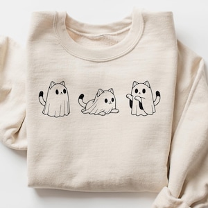 Pu&ograve; includere: Una felpa beige con una grafica nera di tre gatti vestiti da fantasmi. I gatti indossano lenzuola bianche con le code che sporgono.