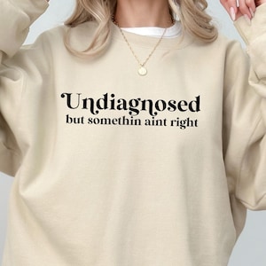 May include: A beige sweatshirt with black text that reads "Undiagnosed but somethin aint right".