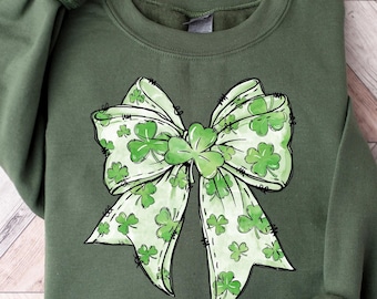 May include: Green sweatshirt featuring a large bow design with shamrocks. The bow is white with green shamrock patterns and a green shamrock in the center. The sweatshirt is a solid green color, perfect for St. Patrick's Day.