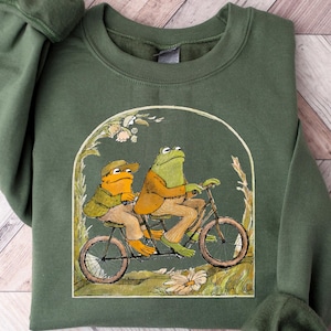 Puede incluir: Una sudadera verde con una ilustración vintage de dos ranas montando una bicicleta. Las ranas llevan sombreros y la bicicleta tiene una cesta en la parte delantera. La ilustración está rodeada por un borde verde.