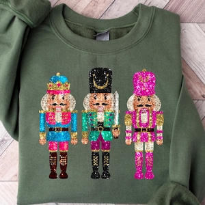 May include: A green sweatshirt featuring a design of three nutcracker soldiers. The nutcrackers are dressed in blue, green, and pink outfits with gold accents and glitter details.