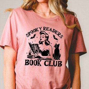 May include: A pink t-shirt with a black graphic of a woman reading a book with a black cat sitting next to her. The graphic says "Spooky Readers Book Club".