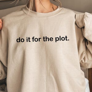 Può includere: Felpa beige con la scritta nera "do it for the plot". La felpa ha un girocollo e maniche lunghe. L'immagine è stata scattata in interni.