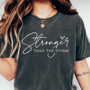 May include: A dark gray t-shirt with the text "Stronger than the storm" in white lettering. The shirt has a relaxed fit and a crew neck.