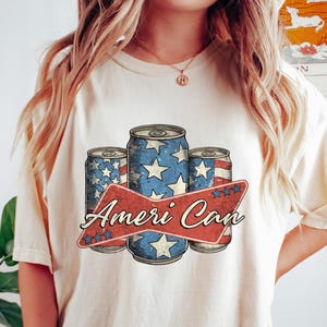 Puede incluir: Camiseta color crema con un gráfico de tres latas de bebidas. Las latas están decoradas con un diseño patriótico, azul con estrellas blancas y rayas rojas. El texto "Ameri Can" está en una pancarta roja.