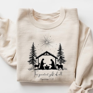 May include: Cream-colored sweatshirt with a black graphic of a nativity scene, including a star, trees, and animals. The text "The greatest gift of all" and Matthew 1:21 are printed below the image. A cozy, long-sleeved top.