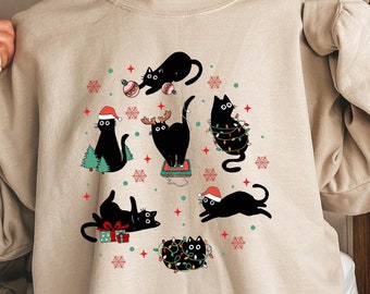 May include: A beige sweatshirt with a Christmas-themed design featuring black cats wearing Santa hats, reindeer antlers, and Christmas lights. The cats are surrounded by snowflakes and red and green baubles.