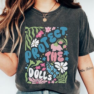 May include: A dark grey t-shirt with a graphic print of colourful flowers and the text "Protect the Dolls".