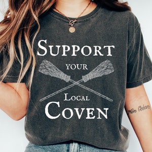 May include: A dark gray t-shirt with a white graphic that says "Support Your Local Coven" with two crossed broomsticks.