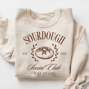 May include: A cream-coloured sweatshirt featuring the words "SOURDOUGH Social Club" in brown lettering. A brown illustration of a loaf of bread is below the word "SOURDOUGH". The sleeves are rolled up.