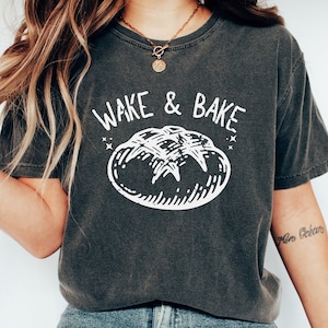 Puede incluir: Una camiseta gris oscuro con un gráfico blanco de una hogaza de pan y el texto "Wake & Bake".