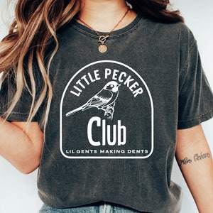 Peut inclure: Un t-shirt gris foncé avec un motif blanc d'un oiseau dans un cercle avec le texte "Little Pecker Club" et "Lil Gents Making Dents".