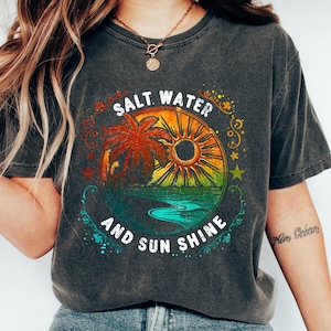 May include: A dark gray t-shirt with a graphic design of a palm tree, sun, and ocean waves. The text "Salt Water And Sun Shine" is written in a circle around the graphic.