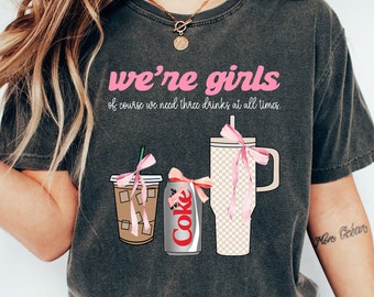 Puede incluir: Camiseta gris oscuro con el texto "we're girls of course we need three drinks at all times". La camiseta presenta ilustraciones de un café helado, una lata de Coca-Cola y un vaso, cada uno adornado con lazos rosas. La camiseta se usa con vaqueros azules y zapatillas blancas.