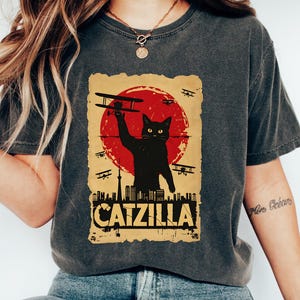May include: Dark gray t-shirt featuring a graphic of a black cat holding a biplane in front of a red sun, with the text "CATZILLA" below. The design includes silhouettes of airplanes and a cityscape.